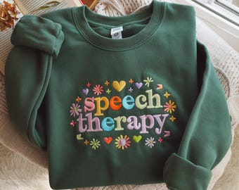 May include: A dark green sweatshirt featuring the embroidered text "speech therapy" in various colours, encircled by floral and heart motifs. The garment is folded on a cream blanket, with a book visible in the background.