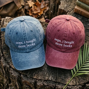 May include: Two baseball caps, one blue and one burgundy, sit side-by-side. Each cap has the phrase "oops, i bought more books" embroidered in white. The caps are on a weathered wooden surface.