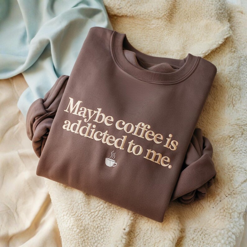 May include: A brown sweatshirt with the embroidered text "Maybe coffee is addicted to me." and a small coffee cup graphic. The sweatshirt is folded on a cream-colored, textured surface.