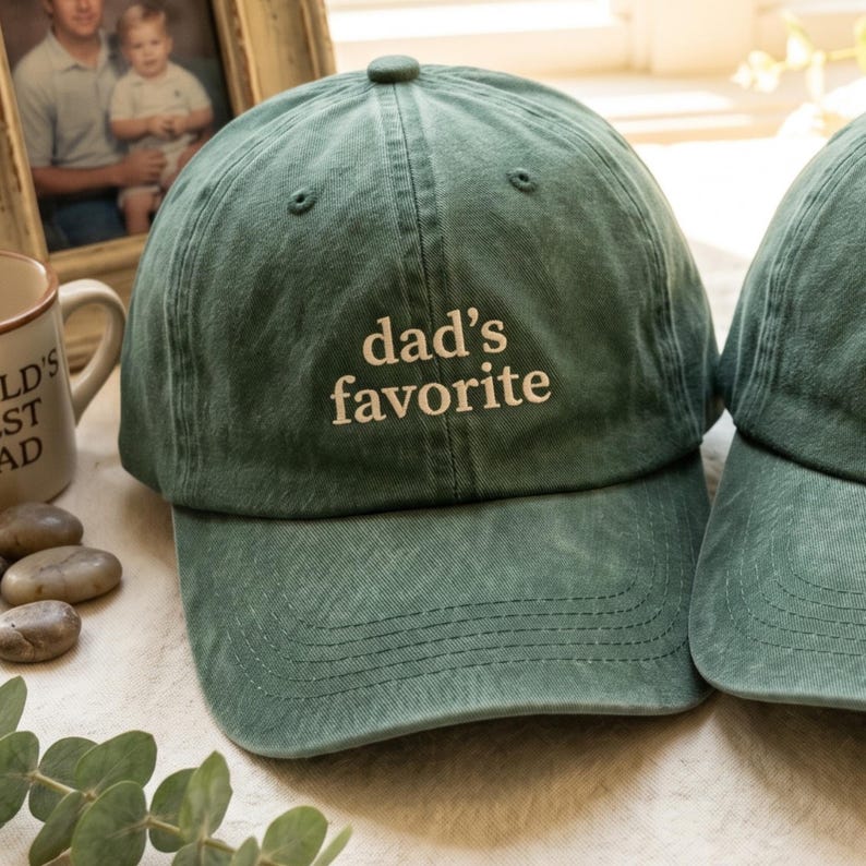 May include: Green baseball cap with the embroidered text "dad's favorite" in white. The cap has a curved brim and a button on top. The cap is made of a soft, textured fabric.