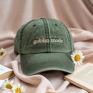 May include: An olive green baseball cap with the words "goblin mode" embroidered in a light pink script. The cap is resting on a soft, peach-colored fabric, with daisies and a book nearby.