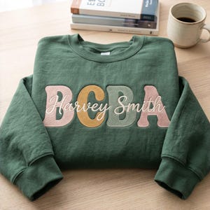 May include: A dark green sweatshirt featuring the embroidered text "BCBA Harvey Smith". The letters are in pink, yellow, and green, with the name in white script. The sweatshirt is folded on a wooden surface.