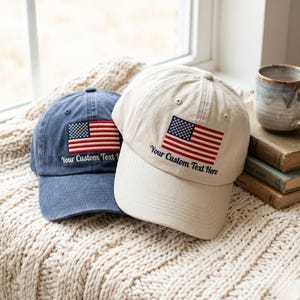 May include: Two baseball caps, one in navy blue and one in cream, each featuring an embroidered American flag patch on the front panel. Below the flag, the text Your Custom Text Here is stitched in dark thread on both caps, which have a structured crown.