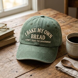 May include: A green baseball cap with the text "I BAKE MY OWN BREAD & DON'T TRUST THE GOVERNMENT" in white. The cap is sitting on a wooden table next to a cup of coffee, a wooden spoon, and a stack of books.