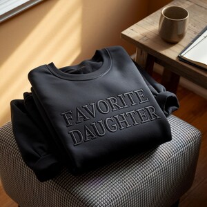 May include: A black sweatshirt with the embroidered text "FAVORITE DAUGHTER" is neatly folded on a patterned ottoman. The sweatshirt is the focal point, with its dark color contrasting against the lighter background. A mug and notebook are visible in the background.