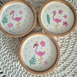 Könnte beinhalten: Drei kreisförmige Untersetzer mit Korkrändern und weißen Zentren. Jeder zeigt eine Illustration eines rosa Flamingos, grüne Blätter, rosa Blumen und gelbe Punkte. Ein Flamingo hat geschlossene Augen, einer offene Augen und einer trägt eine Krone.