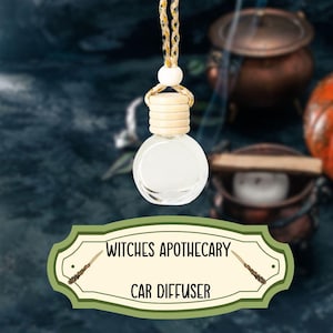 Könnte beinhalten: Eine klare Glas-Autodiffusor-Flasche mit einem Holzdeckel und einem braunen und goldenen Seil, das daran hängt. Die Flasche ist mit "Witches Apothecary Car Diffuser" beschriftet, mit einem Zauberstab auf jeder Seite des Textes.