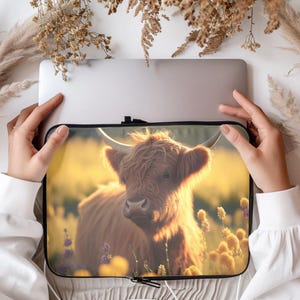 May include: A laptop sleeve featuring a Highland cow in a field of wildflowers. The cow has long, shaggy brown fur and large horns. The background is a soft, golden light, suggesting a sunset or sunrise. The sleeve has a black zipper.