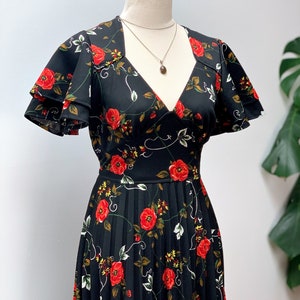 May include: A black maxi dress with a floral print featuring red roses and green leaves. The dress has a V-neckline, short sleeves with ruffles, and a pleated skirt.