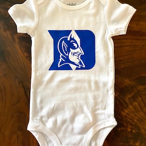 May include: A white short-sleeve baby bodysuit featuring a large blue letter D with a white devil mascot graphic inside the letter. The garment has a round neckline, short sleeves, and three metal snap closures at the bottom crotch area.