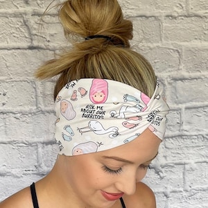 Woman with hair in bun wearing wide twisted headband. The headband is cream color with baby theme print: swaddled babies, baby feet, storks, pacifiers, and "Ask Me About Our Burritos" print throughout.