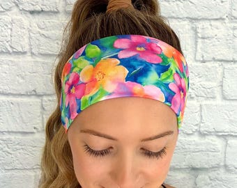 May include: A colorful floral headband featuring a vibrant watercolor design with pink, orange, and yellow flowers on a blue background. The headband is worn by a person with long, light brown hair. The background is a white brick wall.