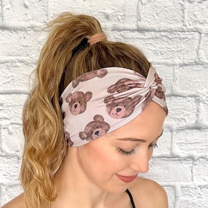 May include: A light pink headband with a repeating pattern of brown teddy bear faces. The headband is twisted at the top and worn on the head. The person in the image has blonde hair pulled back in a ponytail.