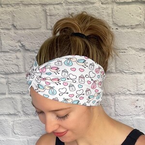 Woman with hair in bun wearing stretchy, wide, twisted headband in white with baby theme print- storks, hearts, bottles, and stethoscopes.