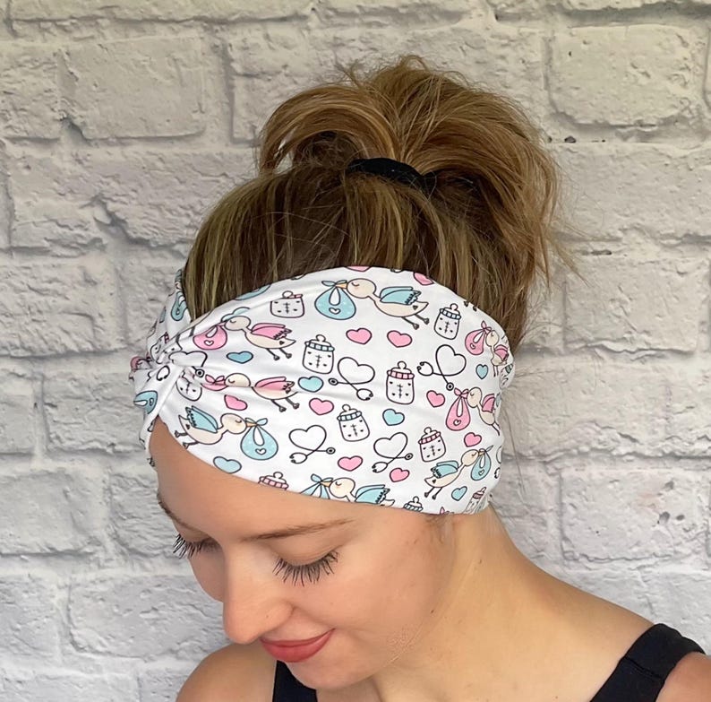 Woman with hair in bun wearing stretchy, wide, twisted headband in white with baby theme print- storks, hearts, bottles, and stethoscopes.