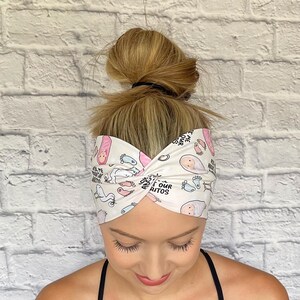 Woman with hair in bun wearing wide twisted headband. The headband is cream color with baby theme print: swaddled babies, baby feet, storks, pacifiers, and "Ask Me About Our Burritos" print throughout.