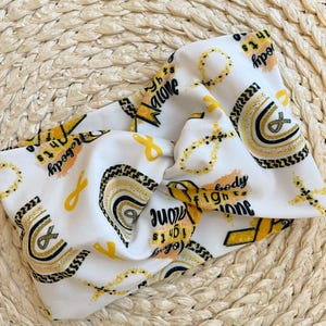 Wide, twisted, white headband with "no one fights alone", gold ribbons, and gold/black rainbow print.