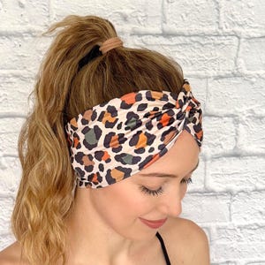 Op de afbeelding: Een luipaardprint hoofdband in bruin, zwart, oranje en groen. De hoofdband is aan de bovenkant gedraaid en wordt over het voorhoofd gedragen. De achtergrond is een witte bakstenen muur.