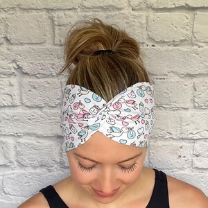 Woman with hair in bun wearing stretchy, wide, twisted headband in white with baby theme print- storks, hearts, bottles, and stethoscopes.