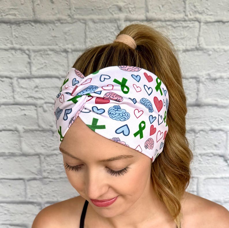Woman with hair in curly ponytail wearing wide, twisted headband with green ribbon, pink and blue brain, and pink and blue heart print.