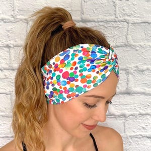 May include: A white headband with a colorful polka dot pattern in shades of red, orange, yellow, green, blue, and purple. The headband is knotted at the top and worn on the head.