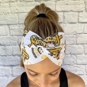 Woman with hair in ponytail wearing wide, twisted, white headband with "no one fights alone", gold ribbons, and gold/black rainbow print.
