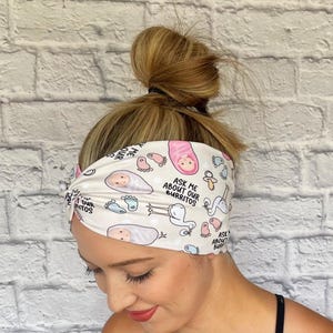 Woman with hair in bun wearing wide twisted headband. The headband is cream color with baby theme print: swaddled babies, baby feet, storks, pacifiers, and "Ask Me About Our Burritos" print throughout.