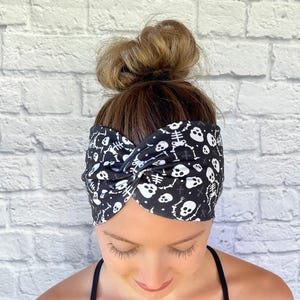May include: A black fabric headband featuring a repeating white pattern of skulls and skeletal rib cages. The headband has a twisted knot detail at the center front and is worn across the forehead, covering the hairline with a wide, fitted band.