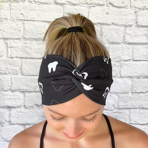 Op de afbeelding: Een zwart hoofdband met een wit tandpatroon. Het hoofdband is gedraaid en wordt gedragen door een persoon met blond haar.