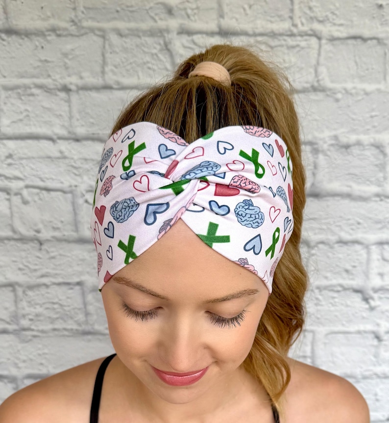 Woman with hair in curly ponytail wearing wide, twisted headband with green ribbon, pink and blue brain, and pink and blue heart print.