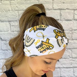 Woman with hair in ponytail wearing wide, twisted, white headband with "no one fights alone", gold ribbons, and gold/black rainbow print.