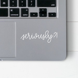 May include: A gray laptop with a white cursive sticker that says "seriously?!" The laptop keyboard is visible, with keys like command, option, and shift. The laptop is on a white wooden surface.