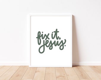 May include: White framed art print with the phrase "fix it. jesus." in a handwritten style, using a dark green color. The artwork is displayed on a white wall above a wooden floor.