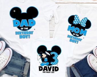 Op de afbeelding: Drie witte T-shirts met blauwe en zwarte Mickey Mouse-ontwerpen. De shirts zeggen "Papa van de jarige jongen!" "Mama van de jarige jongen!" en "David jarige jongen".