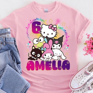 May include: Pink t-shirt featuring a colorful graphic of cartoon characters, the number 6, and the name "Amelia". The shirt is paired with blue jeans and white sneakers. A bouquet of pink flowers is also visible.