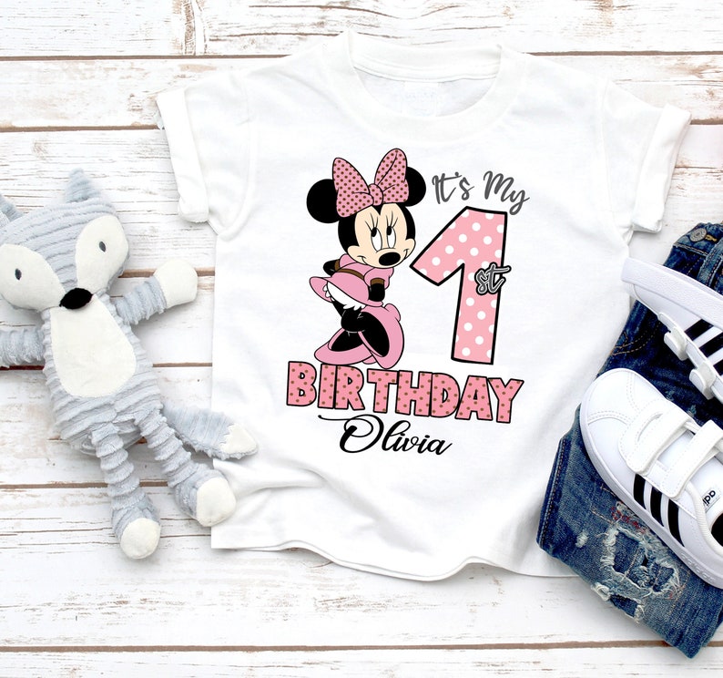 May include: White t-shirt with a pink Minnie Mouse graphic, a pink and white polka dot number one, and the text "It's My 1st Birthday Olivia".