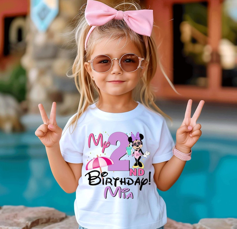 May include: A young child wearing a white t-shirt with a graphic that says "My 2nd Birthday! Mia" and a Minnie Mouse design. The child is wearing a pink bow, sunglasses, and a pink bracelet. The child is holding up two fingers.