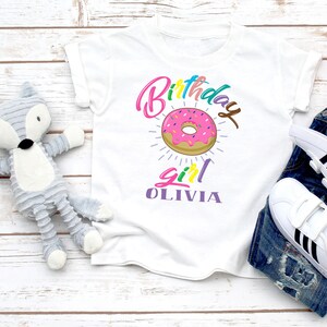 May include: White t-shirt with a colorful "Birthday Girl" graphic featuring a pink and white donut with sprinkles. The name "Olivia" is written in purple below the donut.