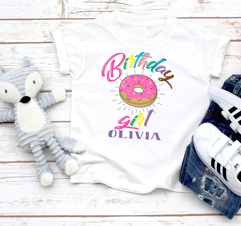 May include: White t-shirt with a colorful "Birthday Girl" graphic featuring a pink and white donut with sprinkles. The name "Olivia" is written in purple below the donut.
