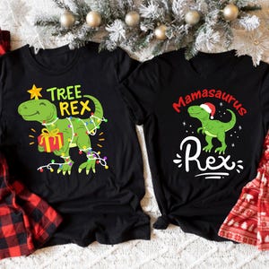Puede incluir: Dos camisetas negras con diseños de dinosaurios con temática navideña. La primera camiseta presenta un dinosaurio verde con una estrella en la cabeza y el texto "Tree Rex". La segunda camiseta presenta un dinosaurio verde con un gorro de Papá Noel y el texto "Mamasaurus Rex".