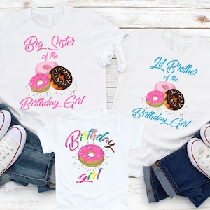 May include: Three white t-shirts with a donut theme. The first shirt says "Big Sister of the Birthday Girl" with pink and brown donuts and sprinkles. The second shirt says "Lil Brother of the Birthday Girl" with pink and brown donuts and sprinkles. The third shirt says "Birthday Girl" with a pink donut and rainbow sprinkles.
