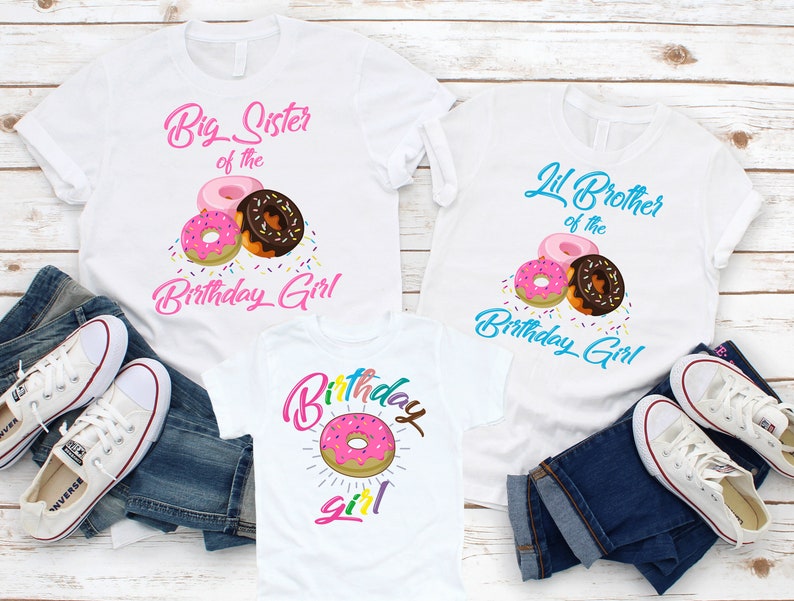 May include: Three white t-shirts with a donut theme. The first shirt says "Big Sister of the Birthday Girl" with pink and brown donuts and sprinkles. The second shirt says "Lil Brother of the Birthday Girl" with pink and brown donuts and sprinkles. The third shirt says "Birthday Girl" with a pink donut and rainbow sprinkles.