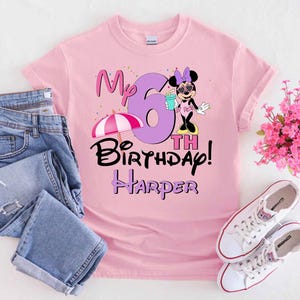 May include: Pale pink t-shirt featuring a Minnie Mouse design, the text "My 6th Birthday! Harper", and a beach umbrella illustration. The shirt is shown with blue jeans and white trainers.