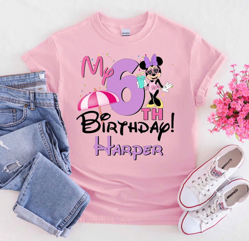 May include: Pale pink t-shirt featuring a Minnie Mouse design, the text "My 6th Birthday! Harper", and a beach umbrella illustration. The shirt is shown with blue jeans and white trainers.