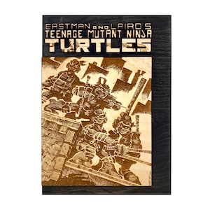 Può includere: Targa in legno con un'immagine incisa al laser delle Tartarughe Ninja. L'opera raffigura le tartarughe in azione, con il testo "EASTMAN AND LAIRD'S TEENAGE MUTANT NINJA TURTLES" in alto. La targa ha una cornice in legno scuro.