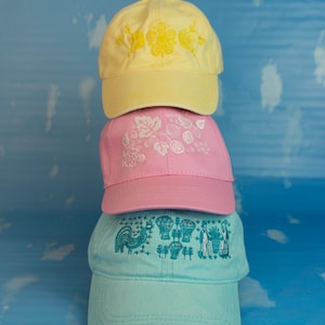 May include: Three baseball caps stacked on top of each other. The top cap is yellow with a floral design, the middle cap is pink with a botanical design, and the bottom cap is light blue with a rooster and other graphics.