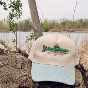 Puede incluir: Una gorra de béisbol con corona color crema y visera de mezclilla azul claro. El panel frontal muestra una trucha arcoíris bordada con detalles en verde, rosa y negro. La gorra presenta un diseño estructurado de cinco paneles.