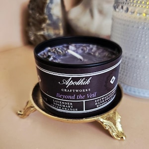 May include: A black candle tin with a white label that reads "Beyond the Veil" and "Apothik Craftworks." The candle sits on a gold-colored decorative stand. The candle's scent includes lavender, rosemary, and sweet orange.