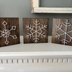 May include: Three wooden blocks with dark brown stain, each featuring a white string art snowflake design. The snowflakes vary slightly in their patterns. These decorative blocks are ideal for winter or holiday decor.