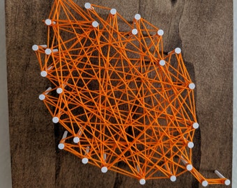 May include: Orange string art on a dark brown wooden background. The string art is in the shape of a leaf with white tacks.